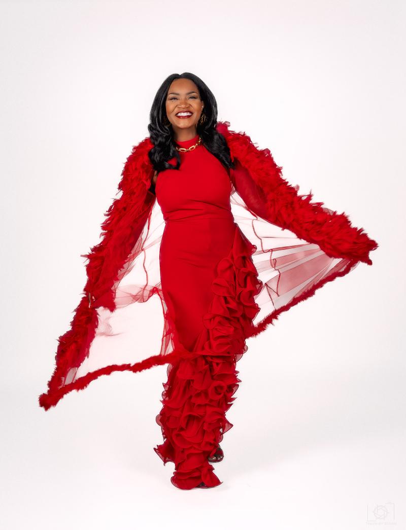 A smiling woman poses in a vibrant red ruffled gown with a flowing feather-trimmed wrap against a white backdrop.