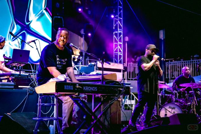 A musician plays a Korg Kronos keyboard on stage illuminated by purple concert lights.