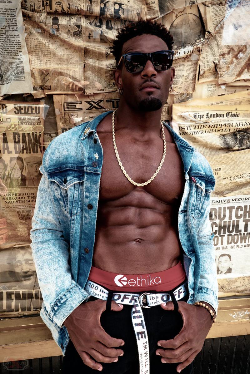 A shirtless man wearing an open denim jacket, gold chain, and sunglasses poses against a wall of vintage newspapers.