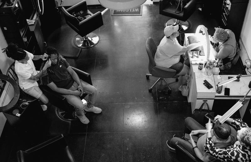 Black and white high-angle view of people sitting in chairs inside a salon on dark tiled floors.