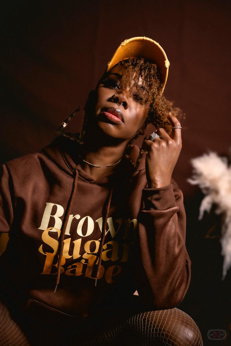 Close-up portrait of a woman wearing a yellow cap and a brown hoodie with gold lettering.