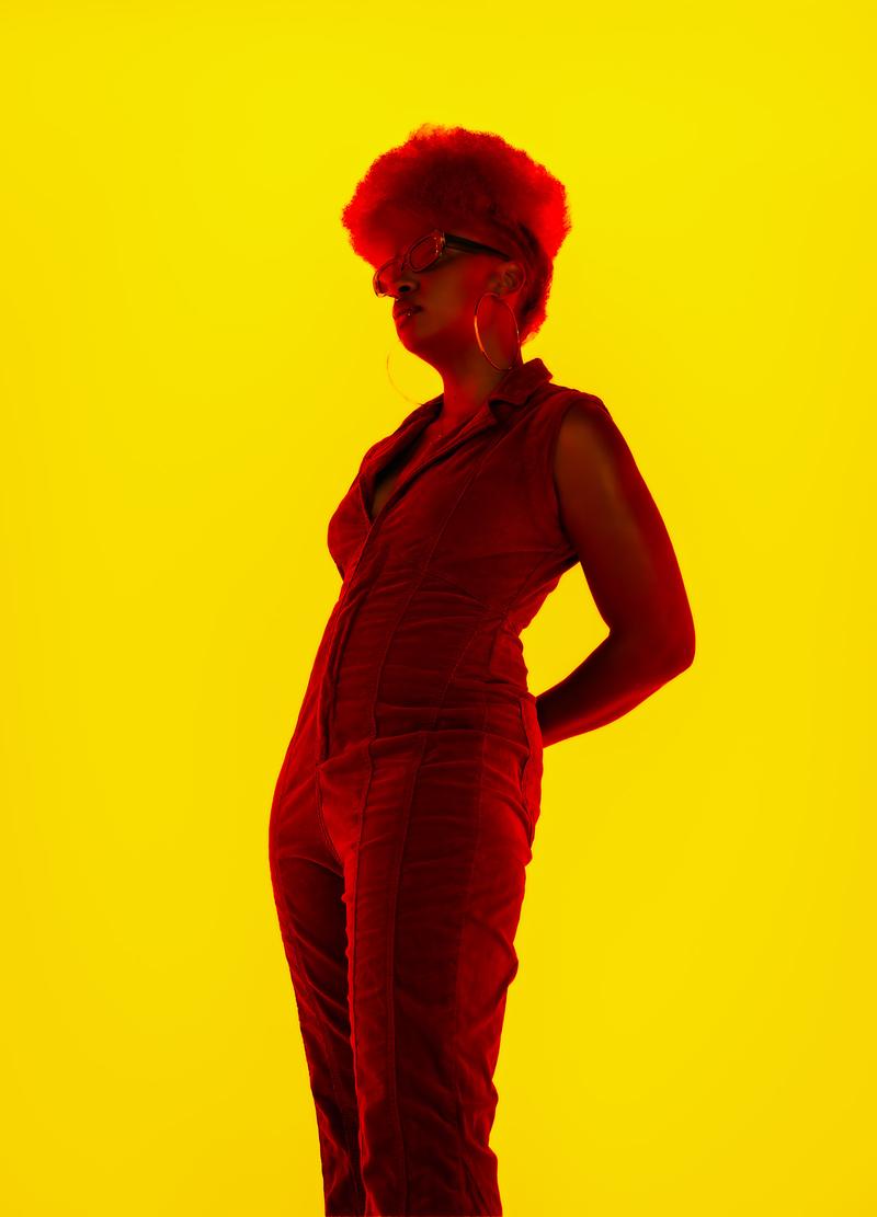 Low-angle studio portrait of a person in a red jumpsuit and sunglasses posed against a bright yellow background.