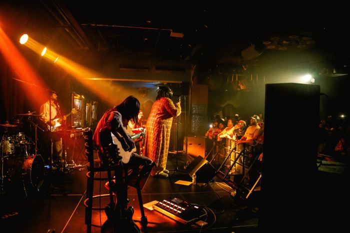 A singer in an orange outfit performs on stage under warm lighting at a live concert venue.