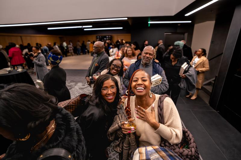 Smiling attendees holding cocktails pose together during a crowded indoor reception event.