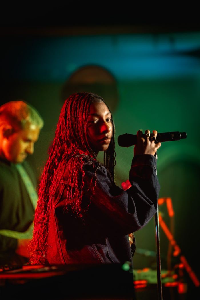 A performer with braided hair holds a microphone under dramatic red and green stage lighting.