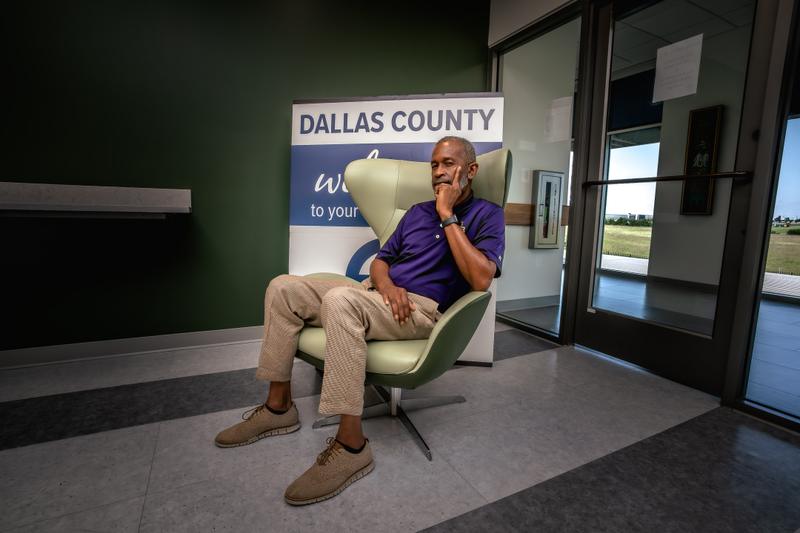 A man in a purple polo shirt sitting thoughtfully in a green armchair in front of a Dallas County sign.