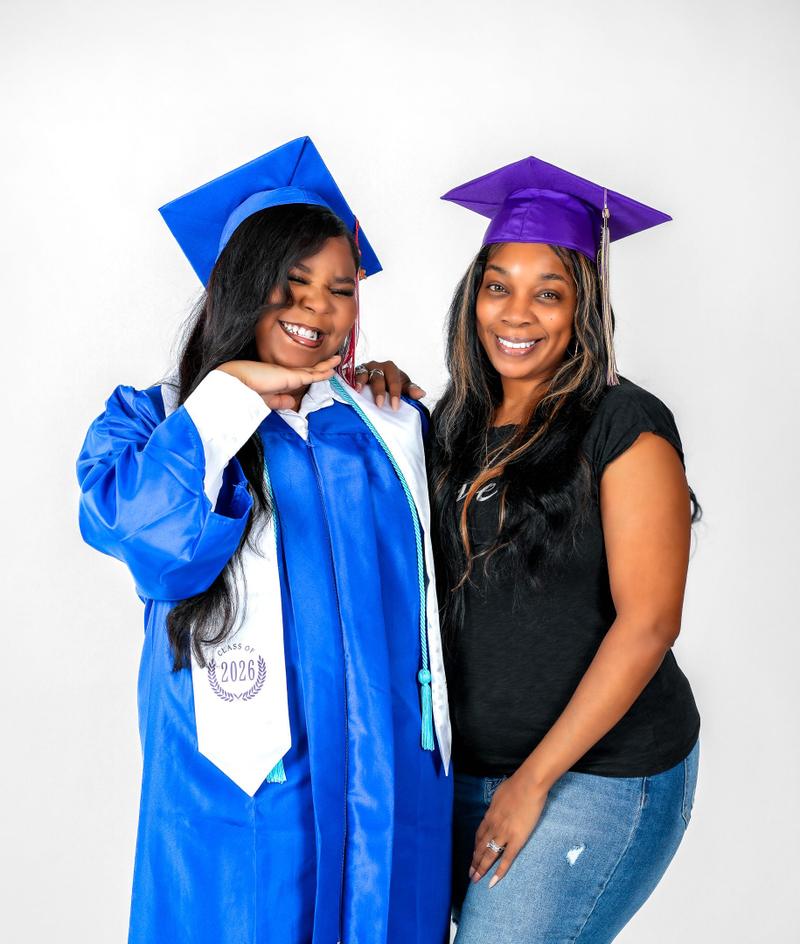 Two women smiling together during a studio graduation photoshoot wearing caps and a Class of 2026 stole.