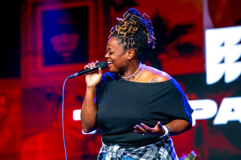 A woman with dreadlocks styled in an updo smiles while singing into a microphone on stage.