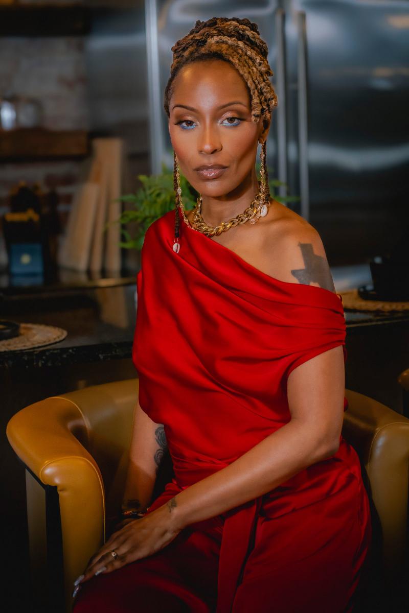 Portrait of a Black woman with styled dreadlocks wearing a red satin top and gold jewelry looking at the camera.