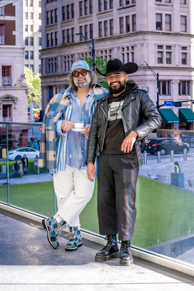 Two stylishly dressed men standing outside in front of a city building, one wearing a black cowboy hat and leather jacket.