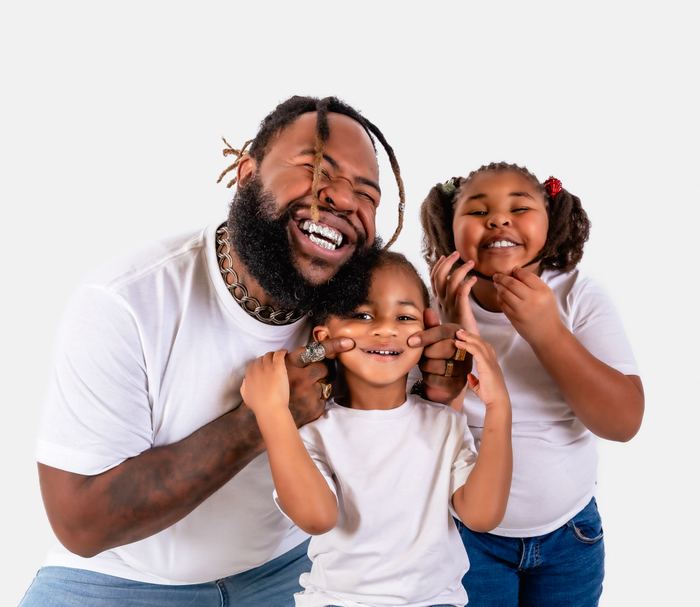 A smiling man with dreadlocks and silver grillz playfully touches a young child's cheeks during a studio photo shoot.