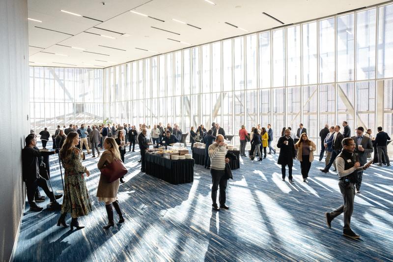 Attendees gather near catering tables inside a modern event hall with tall windows.