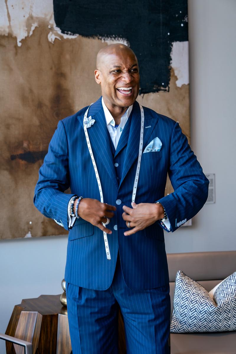 A smiling man wearing a blue pinstripe suit with a measuring tape draped around his neck.