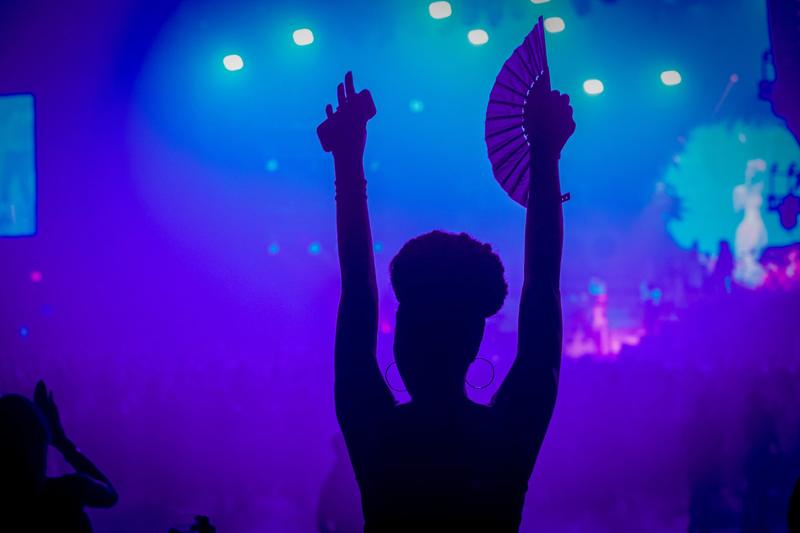 Silhouette of a person in an audience raising their arms with a phone and fan under blue concert lights.