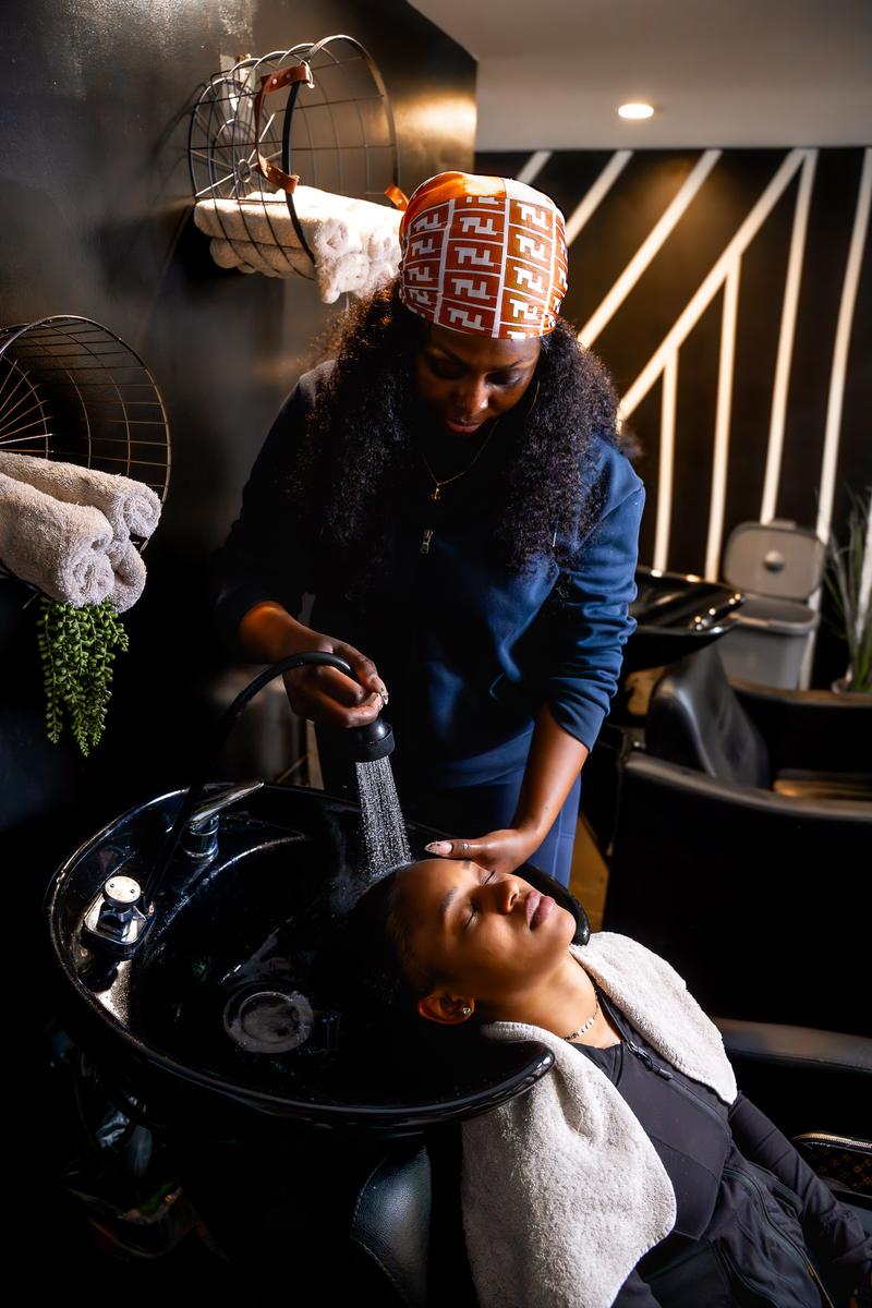 A hair stylist wearing a printed headwrap rinses a client's hair at a salon shampoo basin.