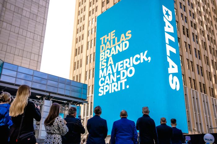 Audience members viewed from behind look up at a large blue banner reading The Dallas Brand Is A Maverick Can-Do Spirit.