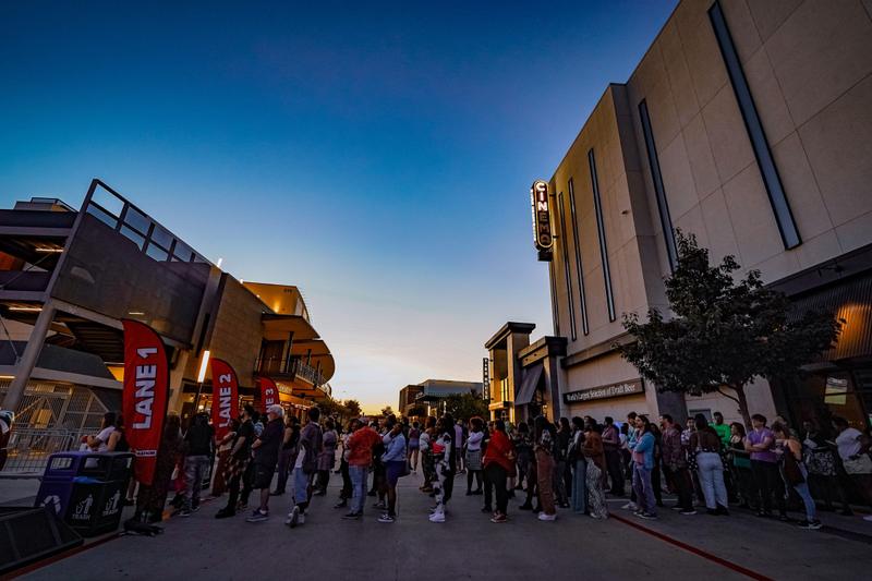 A crowd of people gathers on a sidewalk outside a cinema building at twilight.