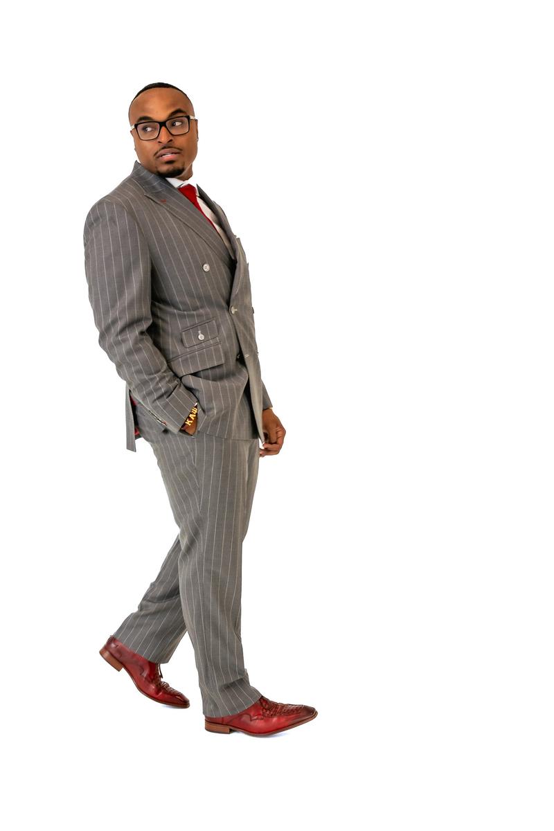 A man wearing a grey pinstripe suit, glasses, and red dress shoes poses against a white background.
