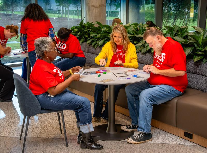 A woman wearing a yellow cardigan and red volunteer shirt works on a craft project at a table during an event.