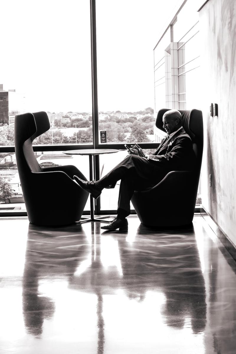 Black and white shot of a man in a suit sitting by a window using his phone in a modern building.
