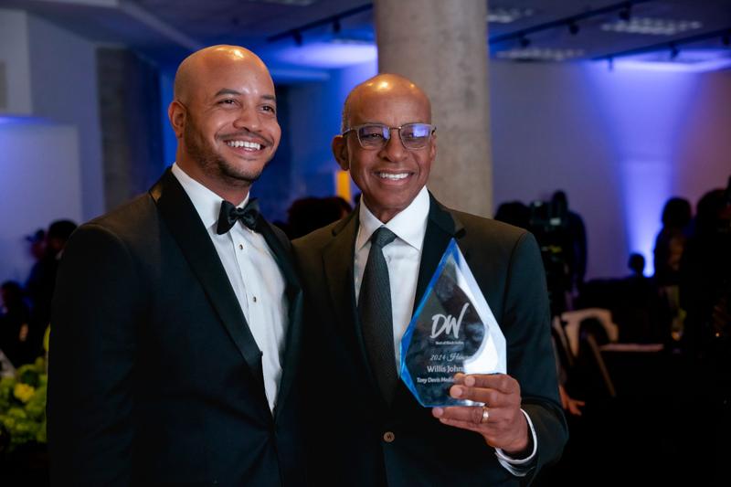 A man in a suit and glasses smiles while holding a glass award plaque at an evening gala.