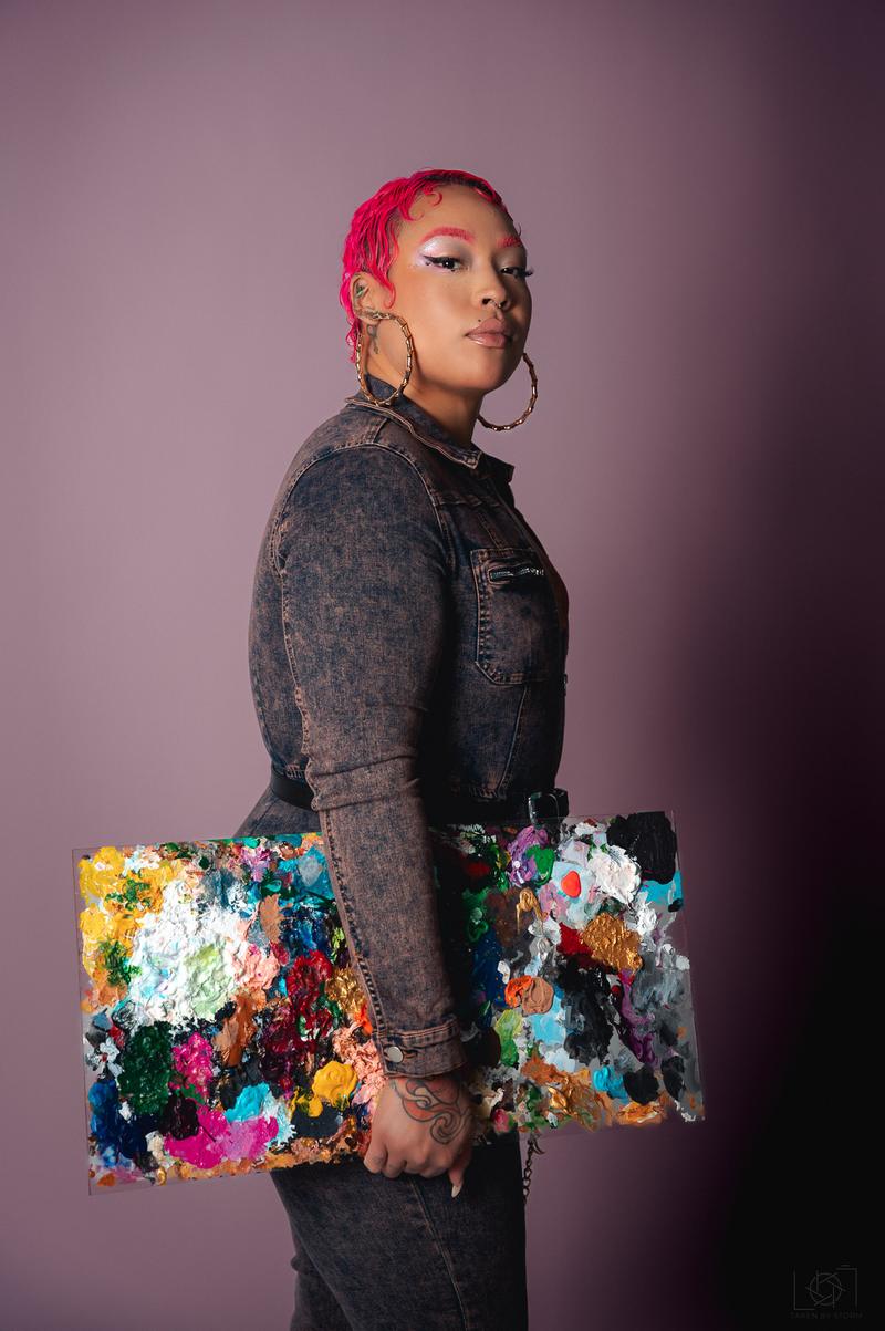 Portrait of an artist with bright pink hair holding a paint-covered clear palette against a studio backdrop.
