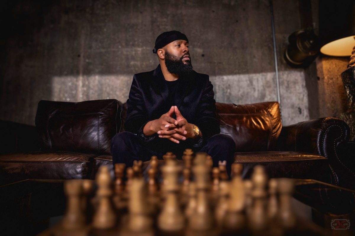A man with a beard sits on a leather couch behind a blurred chessboard in a moody room.