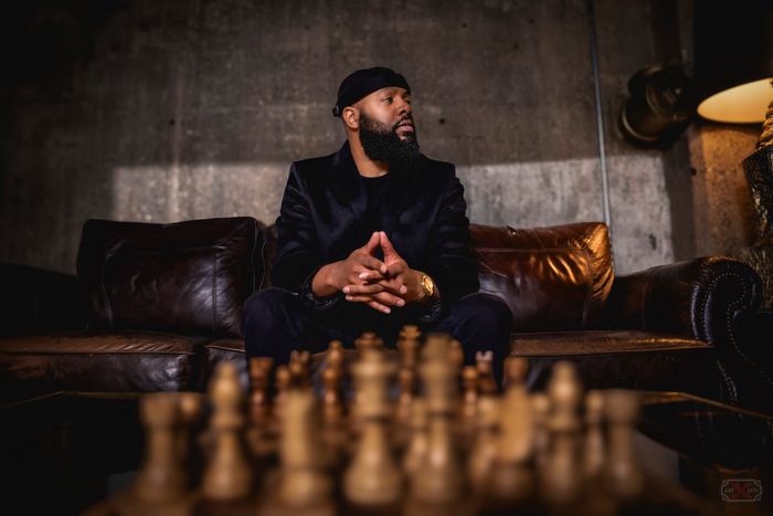 A man with a beard sits on a leather couch behind a blurred chessboard in a moody room.