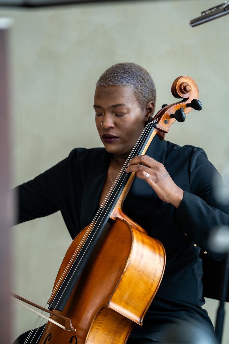 A close-up portrait of a musician with short silver hair playing the cello with eyes closed.
